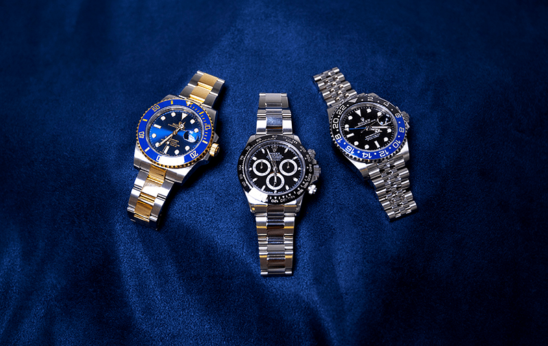 Three luxury wristwatches with metal bands displayed on a dark blue textured surface. The watches vary in dial color and design.