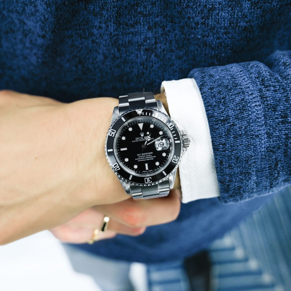 A person wearing a blue sweater and white shirt cuff is showing a silver wristwatch with a black dial on their left wrist.