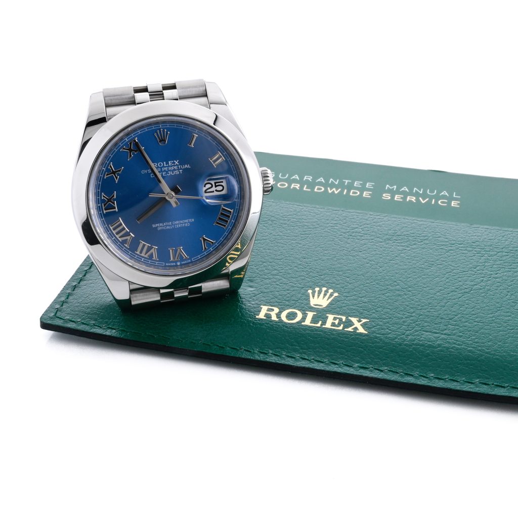 A Rolex wristwatch with a blue dial and Roman numerals is displayed next to a green Rolex guarantee manual.