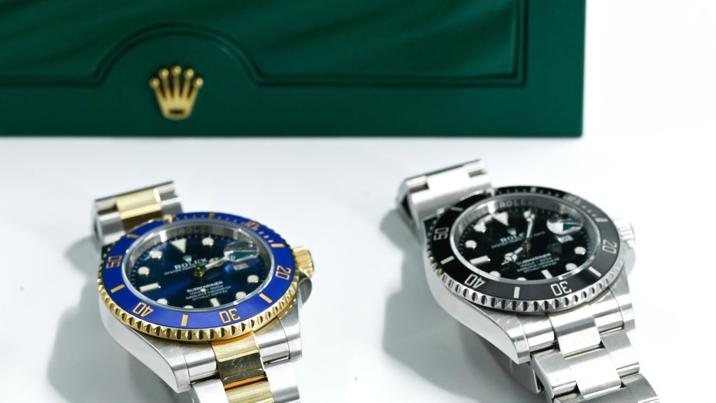 Two Rolex dive watches, each a stunning rolex watchโone with a blue and gold bezel, the other with a black bezelโare displayed in front of a green box adorned with the iconic gold crown logo.