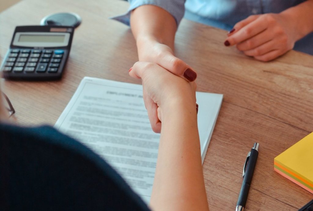 Two people shaking hands over a desk with a contract, a pen, a calculator, and a stack of yellow sticky notes.
