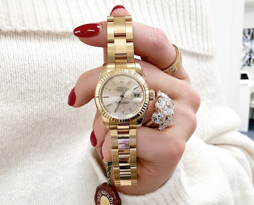 A hand with red painted nails and diamond rings holds a gold Rolex wristwatch with a white dial and date display, perfect for anyone seeking luxury timepieces at Rolex Pawn in Boca Raton.