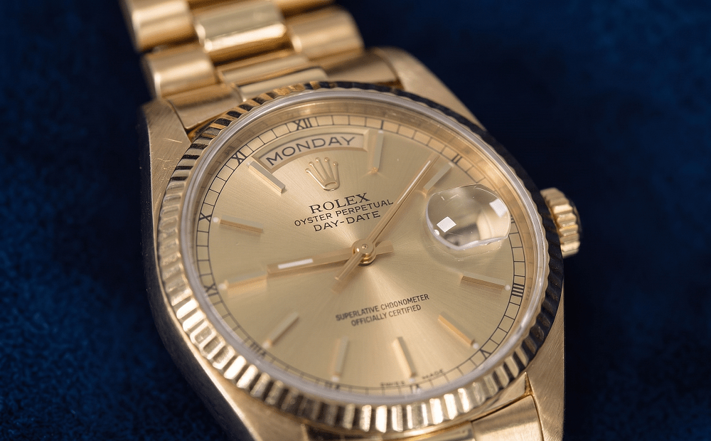 Close-up of a gold Rolex Oyster Perpetual Day-Date watch displaying the day and date, set against a dark blue textured background.